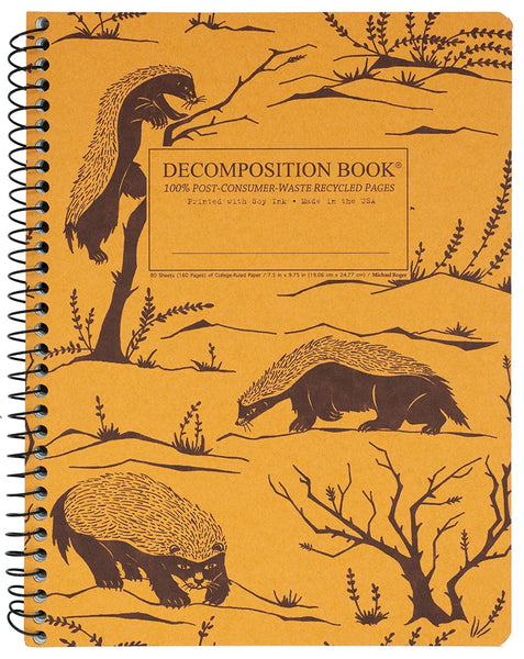 A spiral-bound decomposition book with a yellow-orange cover printed with brown illustrations of honey badgers, trees, and branches. The front cover includes a central label reading 'DECOMPOSITION BOOK' with smaller text below.