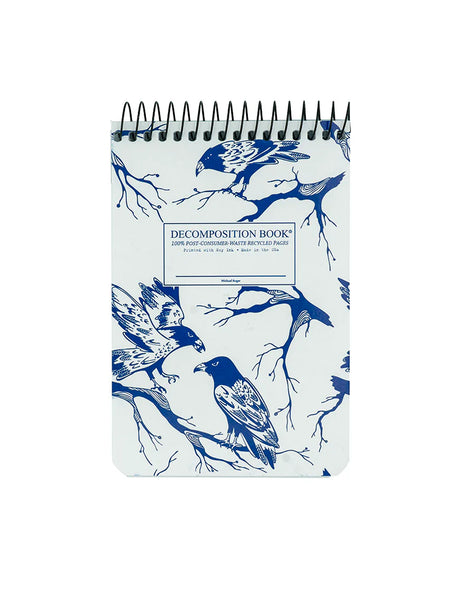 A top spiral-bound decomposition book with a white cover decorated with blue raven birds and branch illustrations, shown on a plain white background. Written in the center, Decomposition Book. 100% Post-Consumer-Waste Recycled Pages. Printed with Soy ink, made in the usa